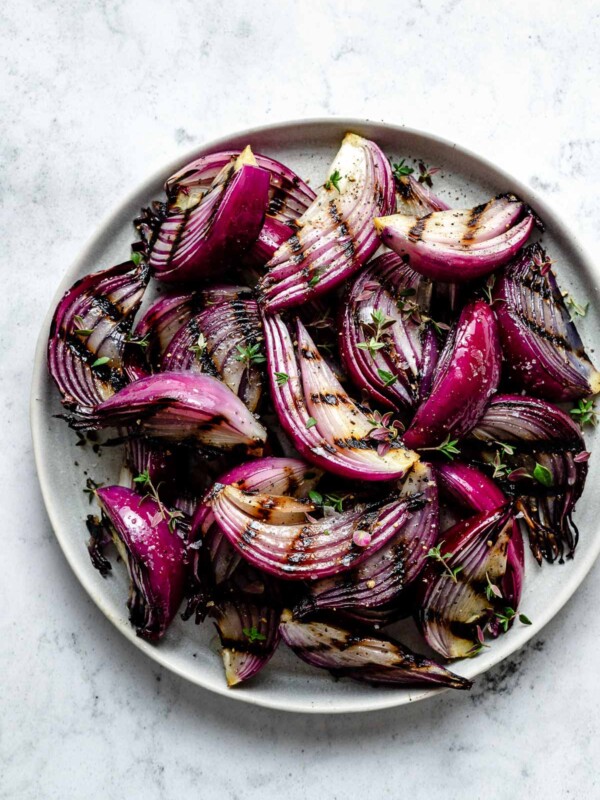 Multiple slices of grilled onion with char marks arranged on a white ceramic plate. The grilled onion is garnished with ground black pepper, kosher salt, & chopped fresh herbs. The plate sits on top of a white & gray marble surface.