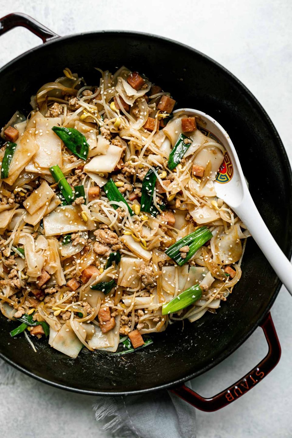 Top down photo of Hawaiian-style pork chow fun in a grenadine colored Staub cast iron wok. The wok sits on a light blue surface with a light blue linen napkin tucked underneath. The chow fun is garnished with sesame seeds and there is a Chinese spoon resting in the dish.