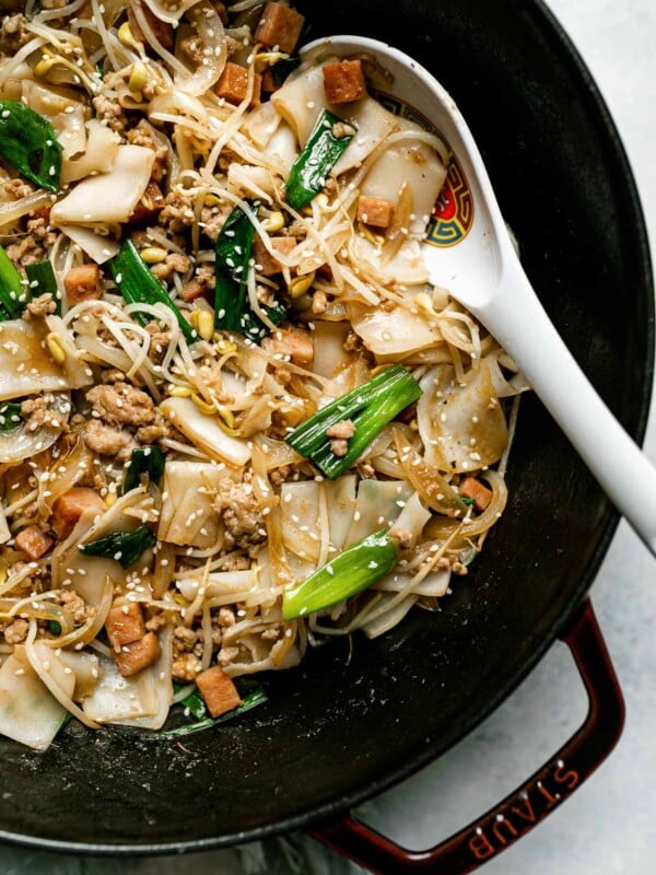 Top down photo of Hawaiian-style pork chow fun in a grenadine colored Staub cast iron wok. The wok sits on a light blue surface with a light blue linen napkin tucked underneath. The chow fun is garnished with sesame seeds and there is a Chinese spoon resting in the dish.