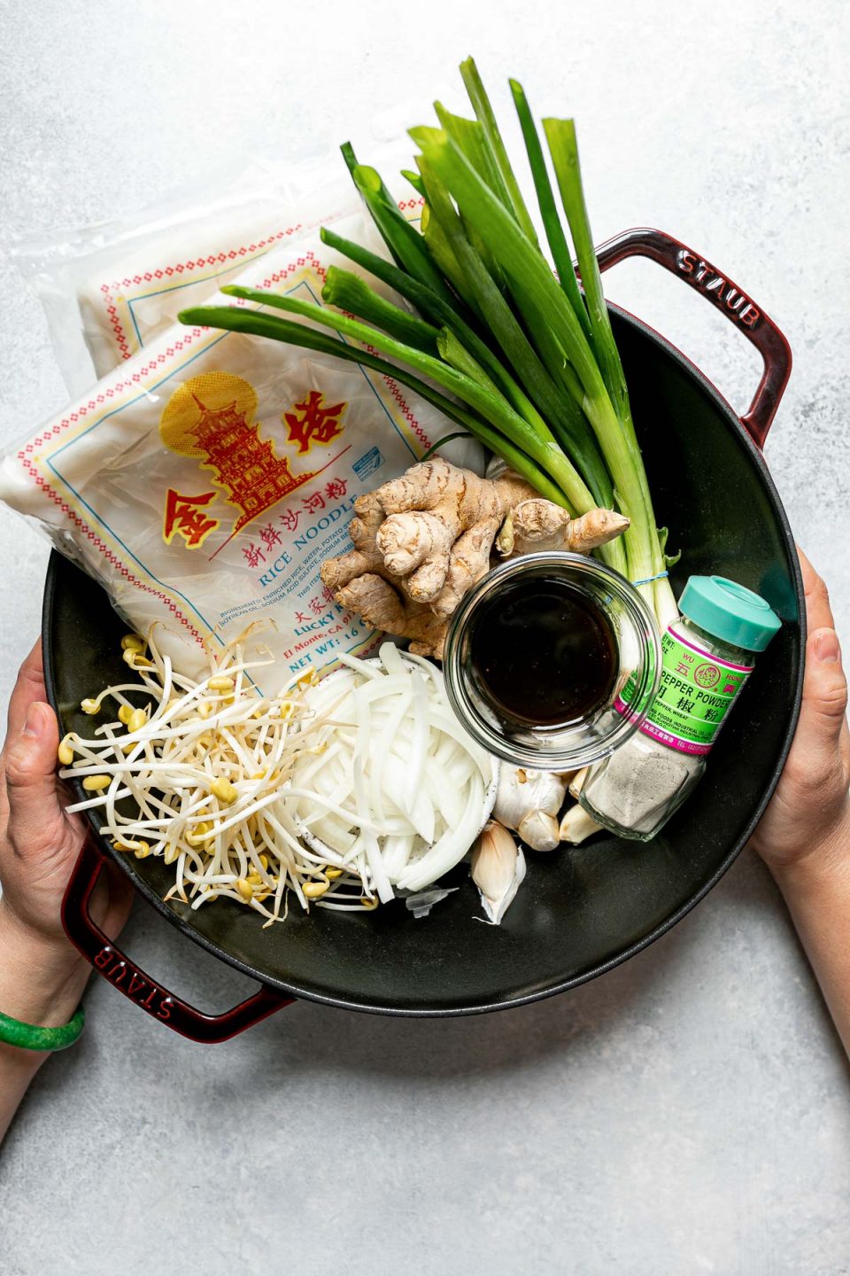 Hawaiian-style pork chow fun ingredients arranged in a grenadine-colored Staub cast iron wok sit on top of a light blue surface: green onions, yellow onions, fresh ginger, garlic, white pepper, fresh rice noodles, bean sprouts, and stir fry sauce that includes oyster sauce, sesame oil, soy sauce, and water. A woman's hands are placed around the wok.