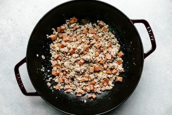 Top down photo of ground pork and diced ham included in pork chow. These ingredients have been cooked inside inside of a grenadine colored Staub cast iron wok. The wok sits on a light blue surface.