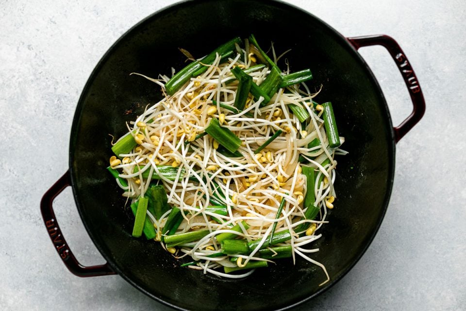 Top down photo of veggies included in pork chow fun including: green onions, yellow onions, and bean sprouts inside of a grenadine colored Staub cast iron wok. The wok sits on a light blue surface.