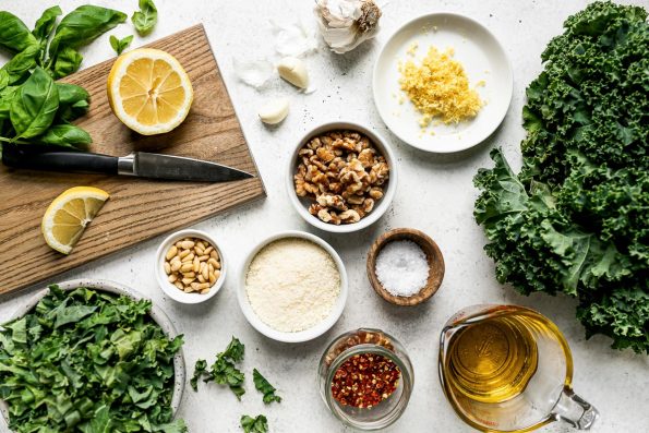 Kale pesto ingredients arranged on a white surface: basil, lemons, kale, pine nuts, walnuts, parmesan, lemon zest, salt, olive oil, red pepper flakes & kale.