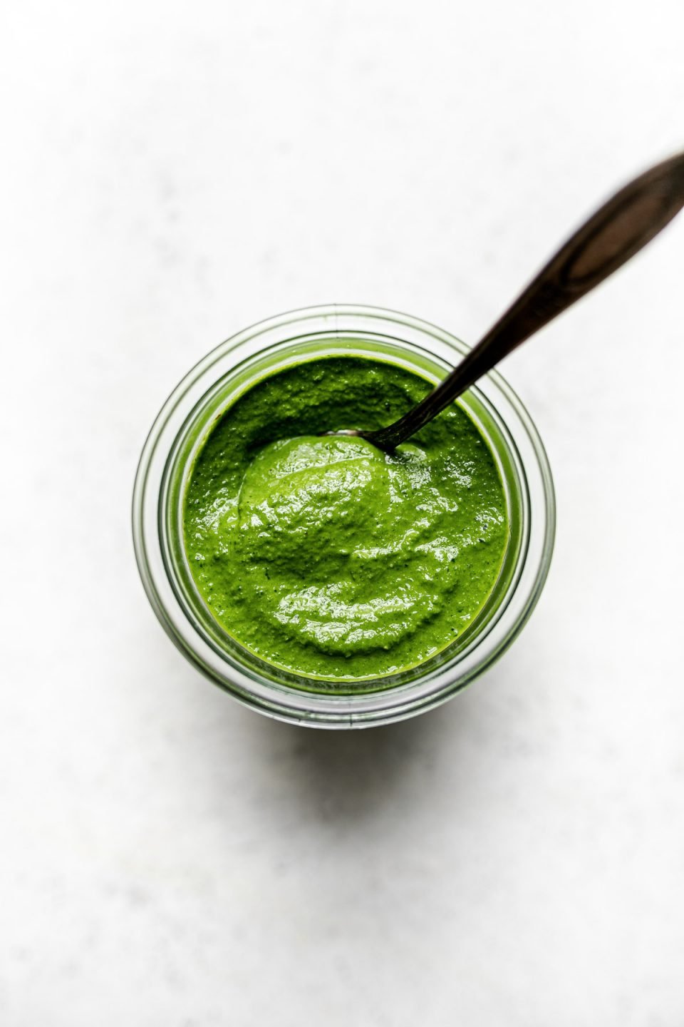 Kale basil pesto in a jar, with a spoon in it. The jar sits atop a white surface.