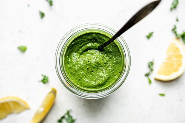 Kale basil pesto in a jar, with a spoon in it. The jar sits atop a white surface surrounded by lemon wedges & shredded kale.