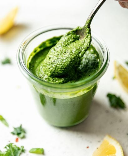 Side angle of woman's hand taking a spoonful of kale pesto out of a jar of kale pesto. The jar sits atop a white surface, surrounded by lemon wedges & shredded kale.