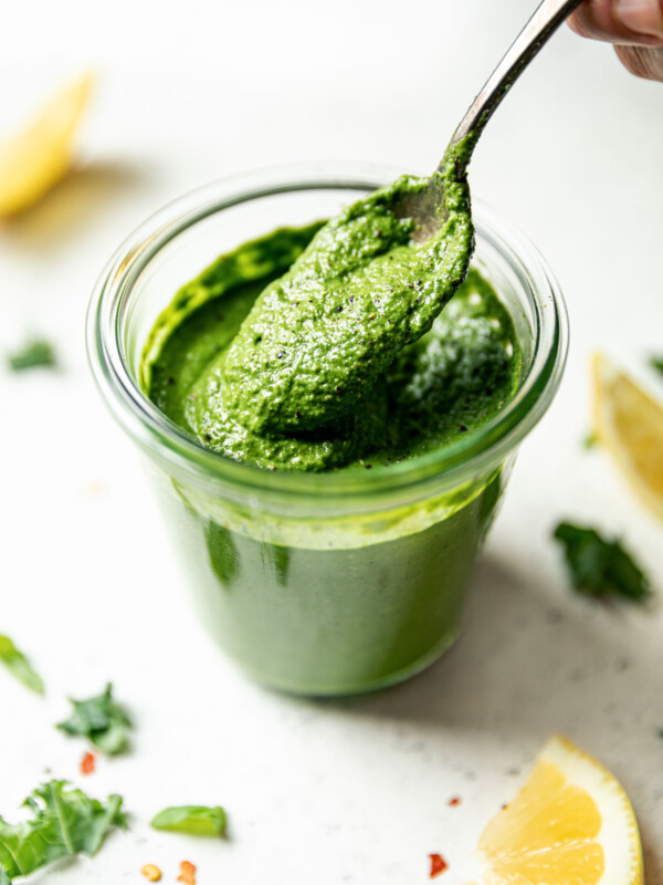 Side angle of woman's hand taking a spoonful of kale pesto out of a jar of kale pesto. The jar sits atop a white surface, surrounded by lemon wedges & shredded kale.