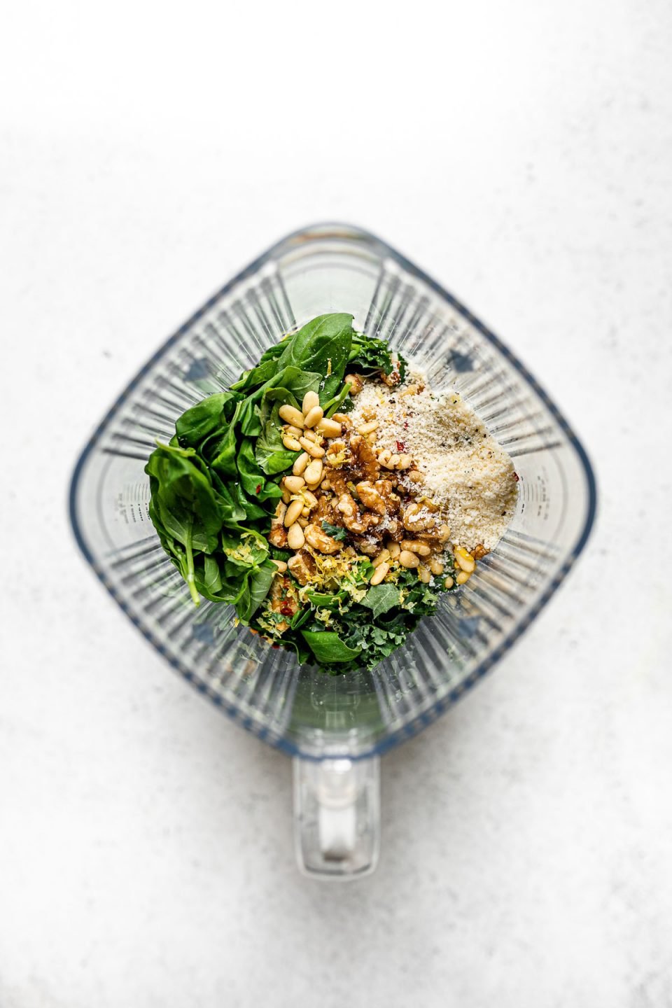 Kale pesto ingredients arranged in a Zwilling Enfinigy Blender – basil, walnuts, parmesan, pine nuts, lemon zest, & kale. The blender jar sits atop a white surface.