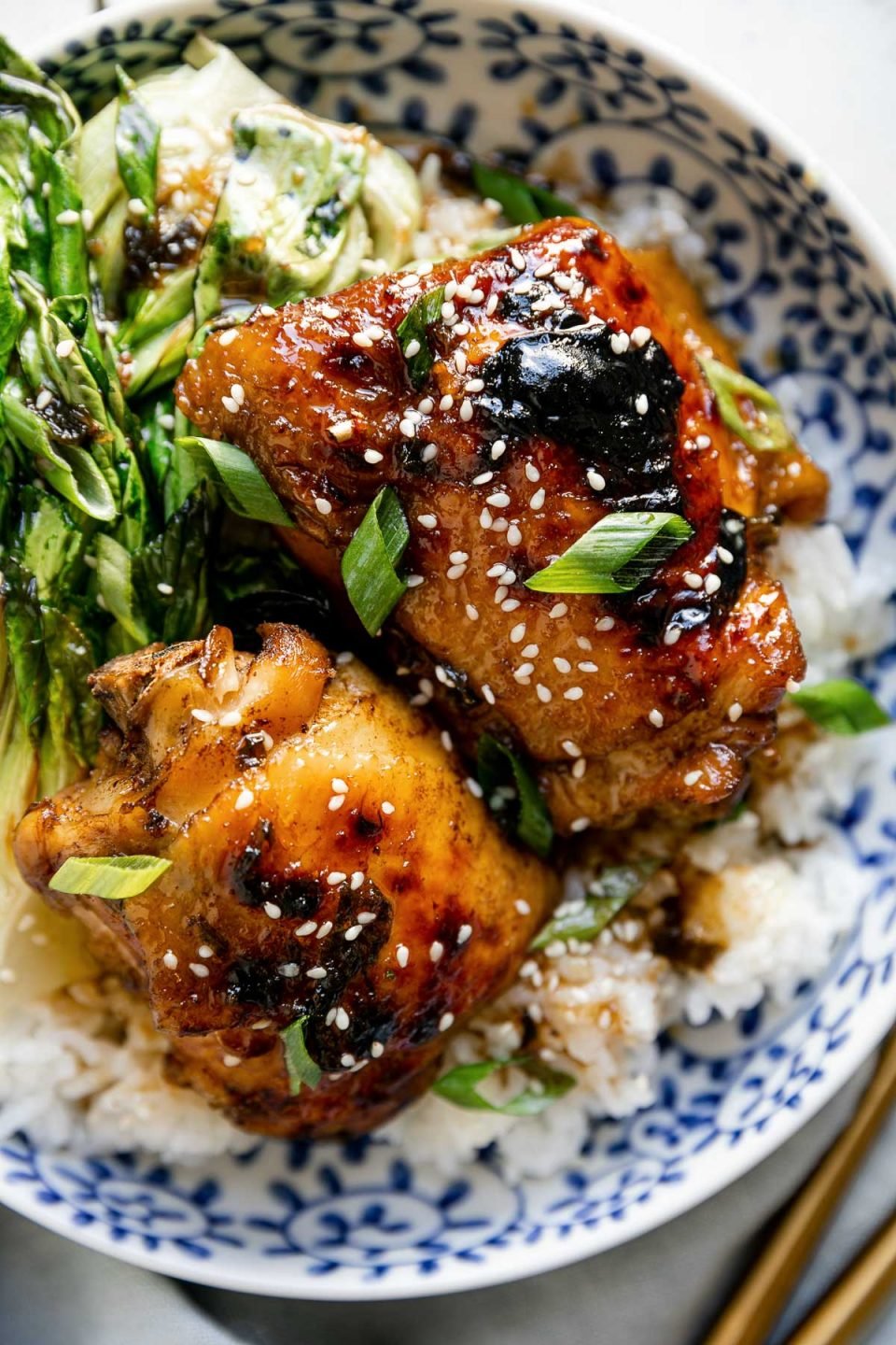 Close up of shoyu chicken plated in a floral-patterened blue dish over rice with bok choy. The chicken is garnished with sliced green onions & sesame seeds. The bowl sits atop a white surface, next to a blue linen napkin & gold flatware.