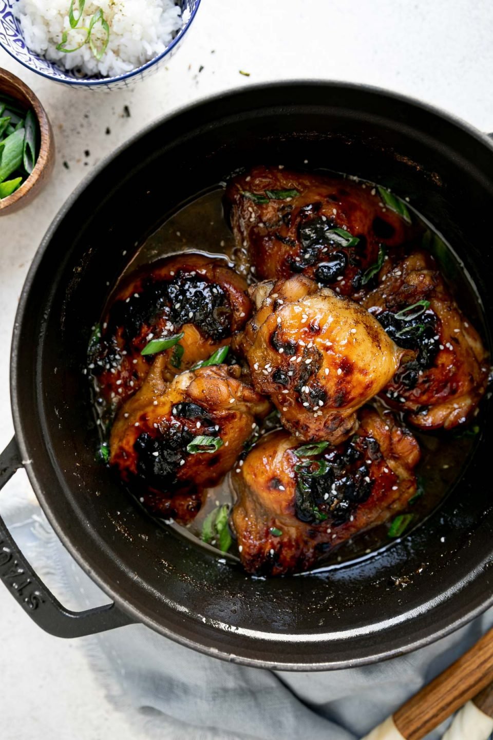 Shoyu chicken in a black dutch oven atop a white surface. The chicken is garnished with sliced green onions & sesame seeds. The dutch oven is surrounded by a blue linen napkin & serving spoons, & 2 small bowls, one filled with rice & the second with sliced green onions.