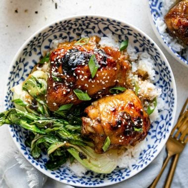 Shoyu chicken plated in a floral-patterened blue dish over rice with bok choy. The chicken is garnished with sliced green onions & sesame seeds. The bowl sits atop a white surface, next to a blue linen napkin, gold flatware, a small bowl of sliced green onion, & a second bowl of shoyu chicken.