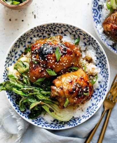 Shoyu chicken plated in a floral-patterened blue dish over rice with bok choy. The chicken is garnished with sliced green onions & sesame seeds. The bowl sits atop a white surface, next to a blue linen napkin, gold flatware, a small bowl of sliced green onion, & a second bowl of shoyu chicken.