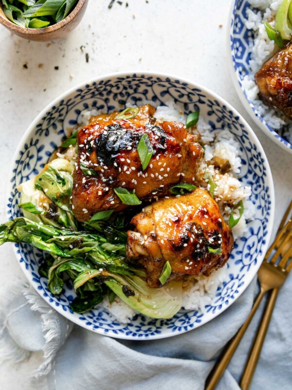 Shoyu chicken plated in a floral-patterened blue dish over rice with bok choy. The chicken is garnished with sliced green onions & sesame seeds. The bowl sits atop a white surface, next to a blue linen napkin, gold flatware, a small bowl of sliced green onion, & a second bowl of shoyu chicken.