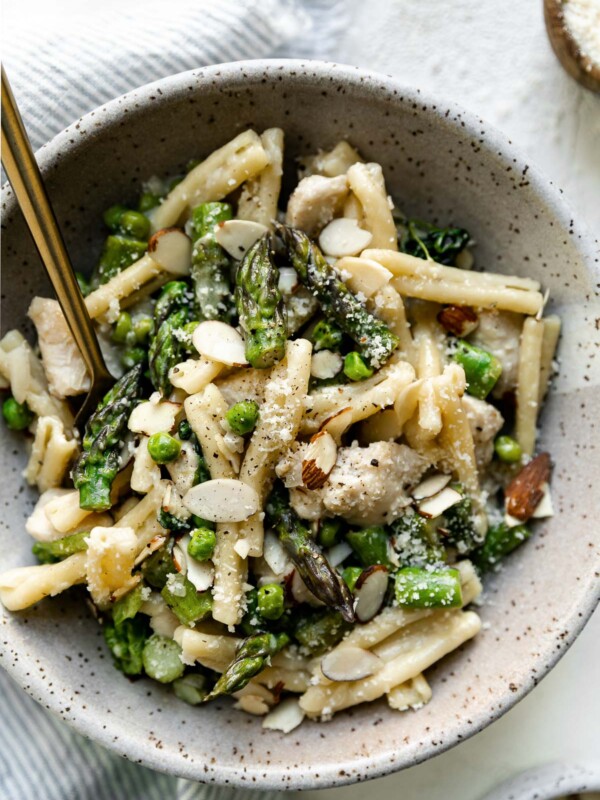 Mascarpone pasta shown in a speckled ceramic bowl, topped with sliced almonds and parmesan cheese. The bowl sits atop a white surface with a gold fork nestled into the pasta, next to a striped blue linen napkin & a small bowl of grated parmesan.