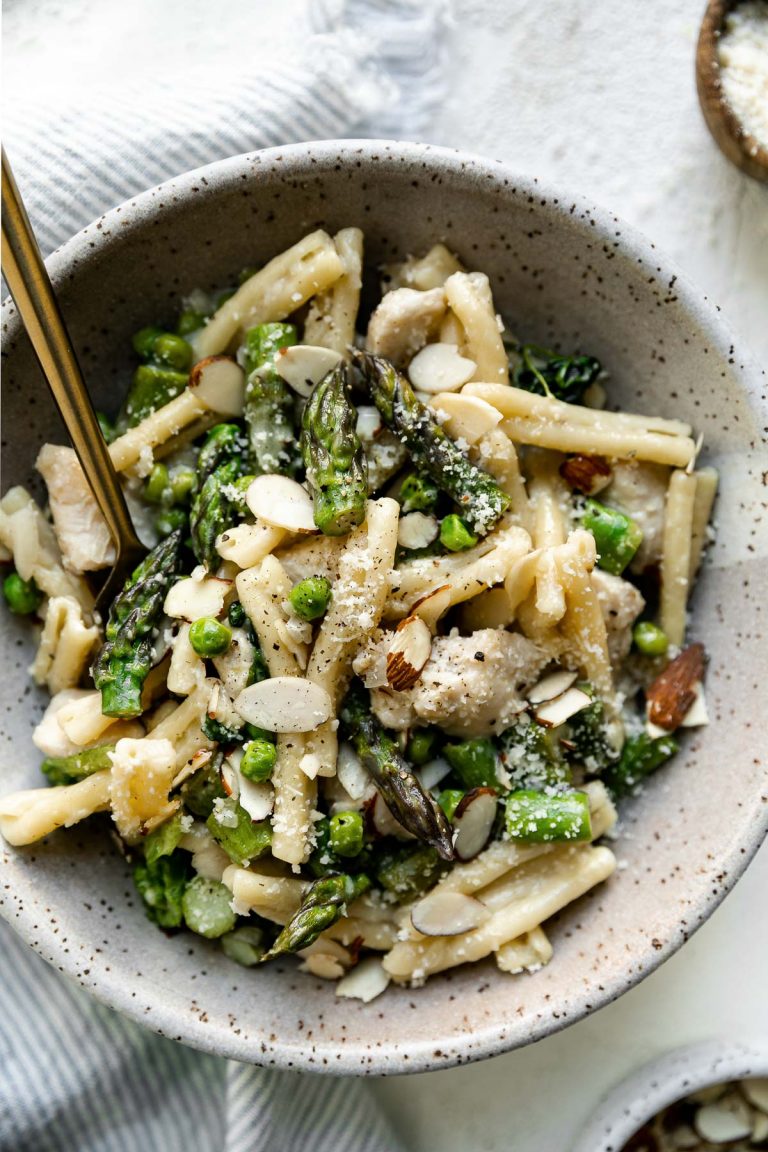 Mascarpone pasta shown in a speckled ceramic bowl, topped with sliced almonds and parmesan cheese. The bowl sits atop a white surface with a gold fork nestled into the pasta, next to a striped blue linen napkin & a small bowl of grated parmesan.