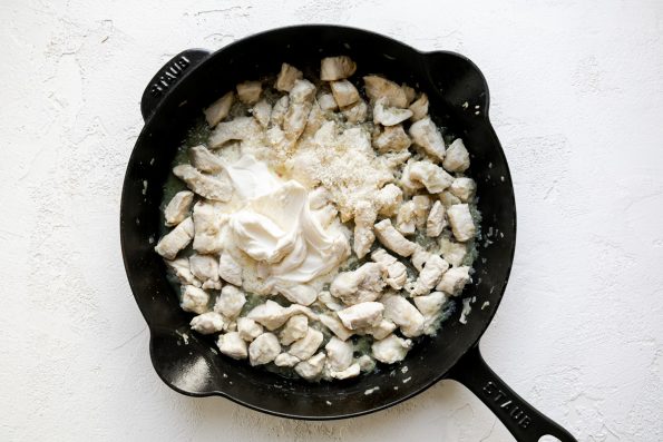 How to make mascarpone pasta sauce, Step 2: Mascarpone & parmesan cheeses on top of browned diced chicken breast & softened shallots in a black cast iron skillet, sitting atop a white surface.