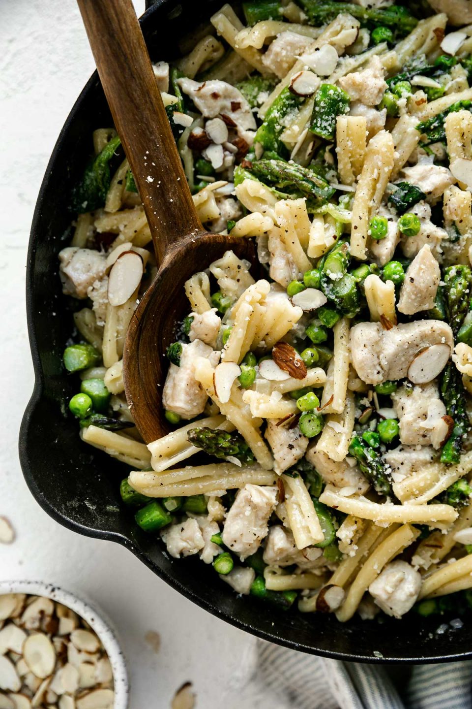 Close up of chicken asparagus pasta in a large black cast iron skillet, atop a white surface. There is a wooden serving spoon nestled in the pasta. The skillet is surrounded by a blue & white striped linen napkin & a small dish of sliced almonds.
