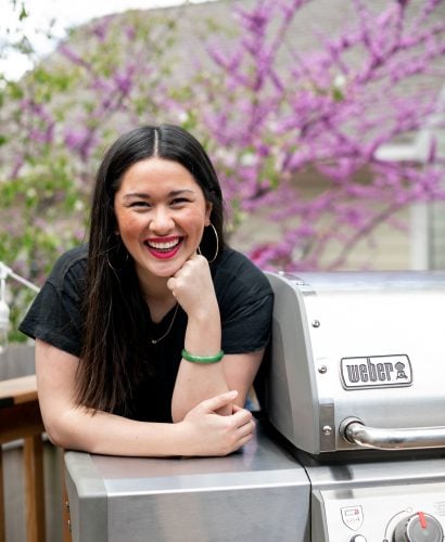 Jess of Plays Well With Butter stands on a deck behind a stainless steel Weber Genesis II propane grill. In the background, a blossoming tree & a house with beige siding.