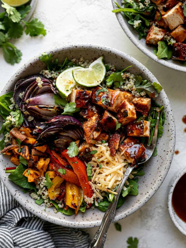 Grilled BBQ chicken bowl with diced BBQ chicken, grilled onions, sweet potatoes & bell peppers sitting atop a bed of greens & quinoa in a large speckled gray bowl. The bowl sits atop a white surface, surrounded by a striped gray linen napkin, a second BBQ chicken rice bowl, extra BBQ sauce, cilantro leaves & lime wedges.