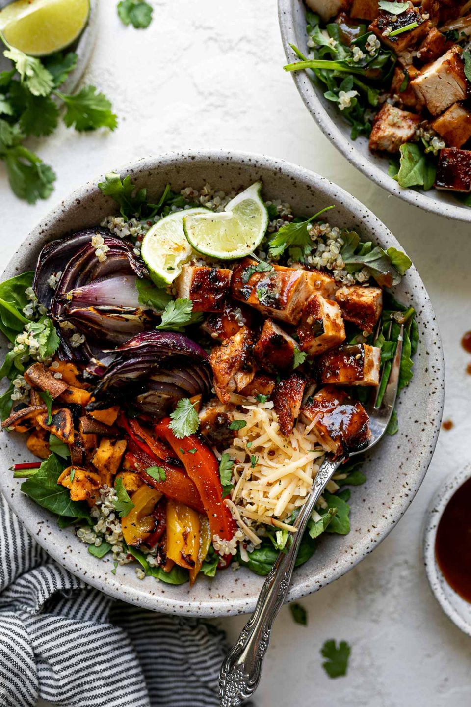 Grilled BBQ chicken bowl with diced BBQ chicken, grilled onions, sweet potatoes & bell peppers sitting atop a bed of greens & quinoa in a large speckled gray bowl. The bowl sits atop a white surface, surrounded by a striped gray linen napkin, a second BBQ chicken rice bowl, extra BBQ sauce, cilantro leaves & lime wedges.