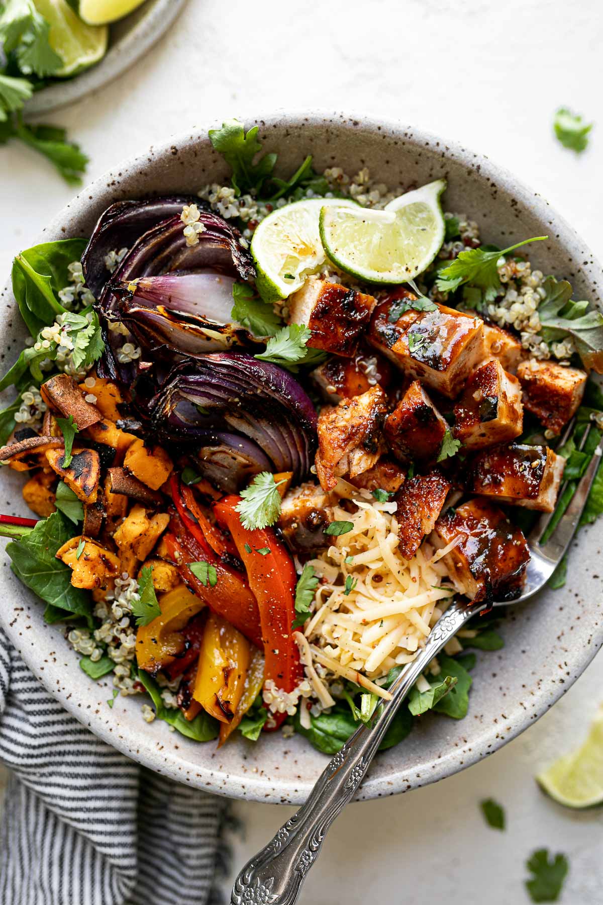 Grilled BBQ chicken bowl with diced BBQ chicken, grilled onions, sweet potatoes & bell peppers sitting atop a bed of greens & quinoa in a large speckled gray bowl. The bowl sits atop a white surface, surrounded by a striped gray linen napkin, cilantro leaves & lime wedges.