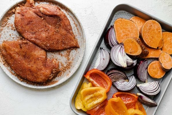 How to make BBQ Chicken Bowls, Step 3: BBQ dry rub-dusted chicken breasts & seasoned sweet potatoes, onions, & bell peppers on a quarter sheet pan atop a white surface.