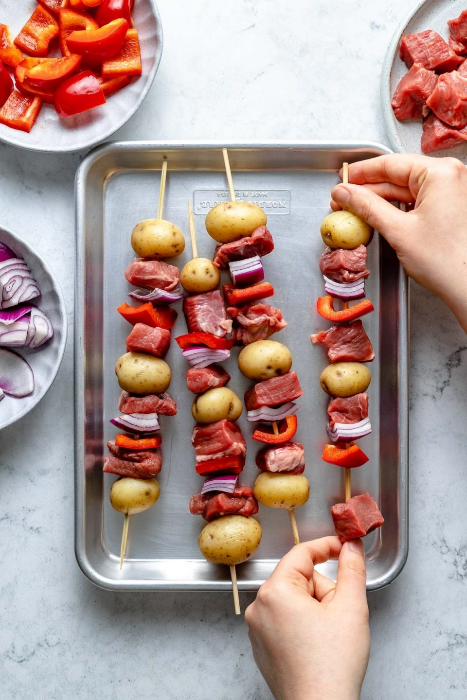 Chimichurri steak and potato kabobs assembly: A woman's hands shown threading steak onto a bamboo skewer with bell peppers, onions, & potatoes. 3 assembled skewers sit atop a small baking sheet on a light blue marbled surface.