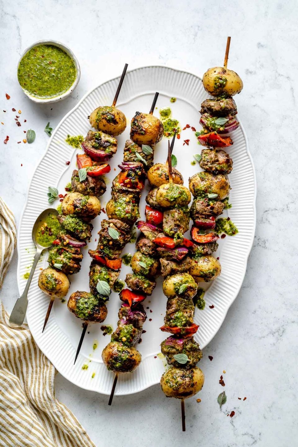 Grilled steak kabobs on a large white oval serving dish, drizzled with chimichurri sauce. The dish sits atop a light blue marbled surface, alongside a small dish of chimichurri sauce & a gold & white striped linen napkin.