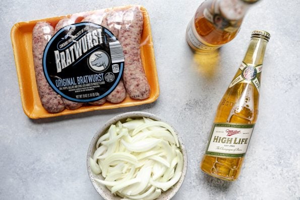 Grilled beer brats ingredients arranged on a light blue surface: Bratwurst from ALDI, Miller High Life, & a bowl of thinly sliced onion.