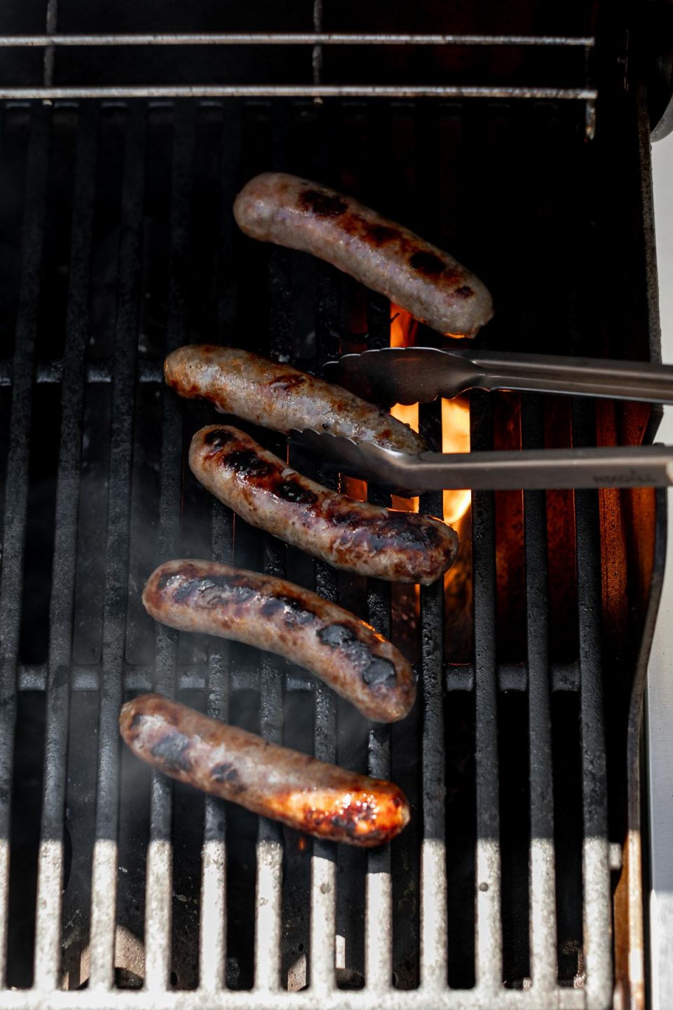 Grilled beer brats over direct heat, charring on grill grates. Grilling tongs peek into the frame, flipping one of the brats.