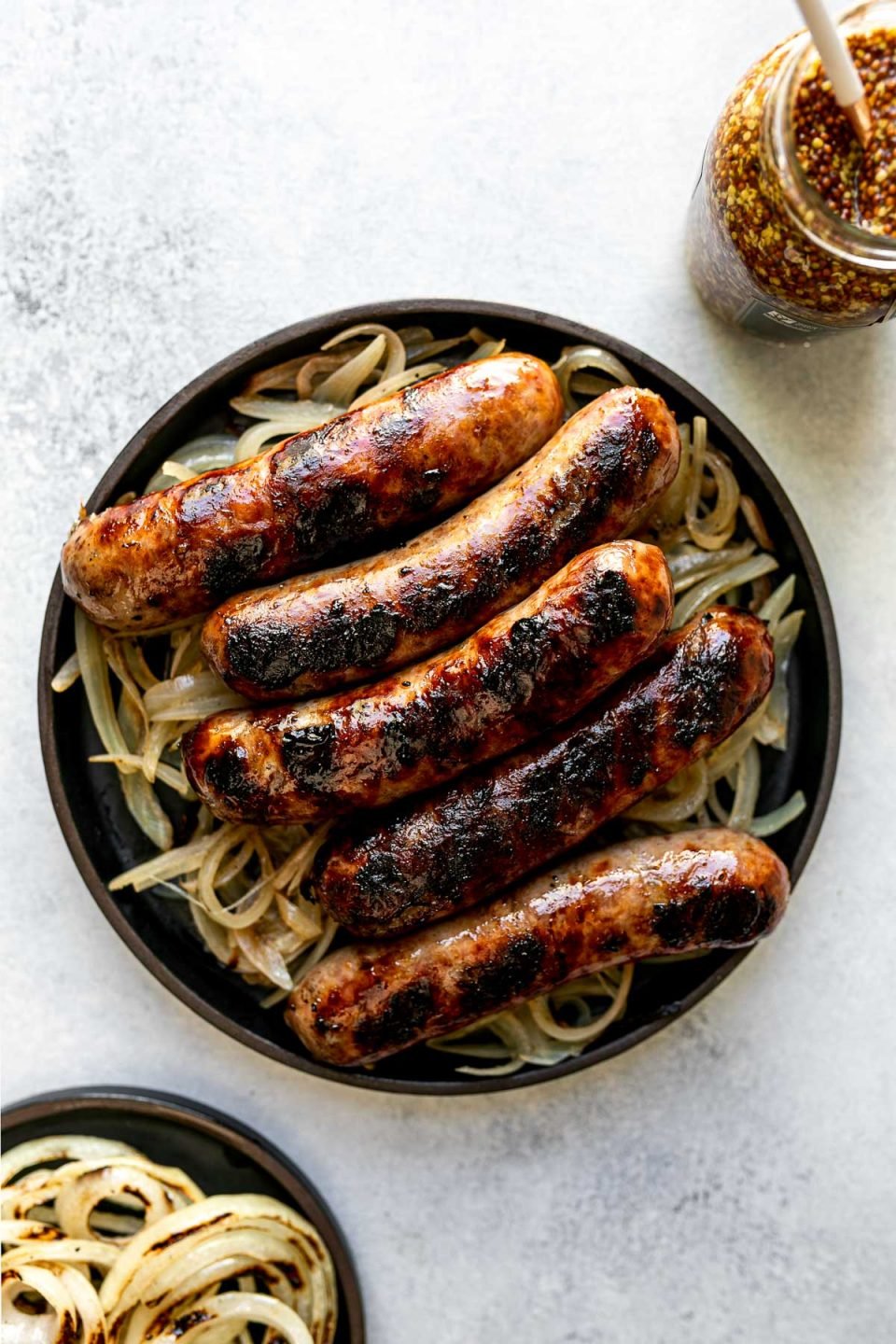 Grilled beer brats sit atop beer braised onions on a black plate. The plate sits atop a light blue surface, next to a small black plate topped with grilled onions & a jar of whole grain mustard.