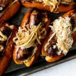Grilled Wisconsin Beer Brats arranged on a small Nordicware baking sheet. The brats sit on toasted brioche buns, topped with mayonnaise, mustard, braised onions, sauerkraut, etc. The baking sheet sits atop a light blue surface.