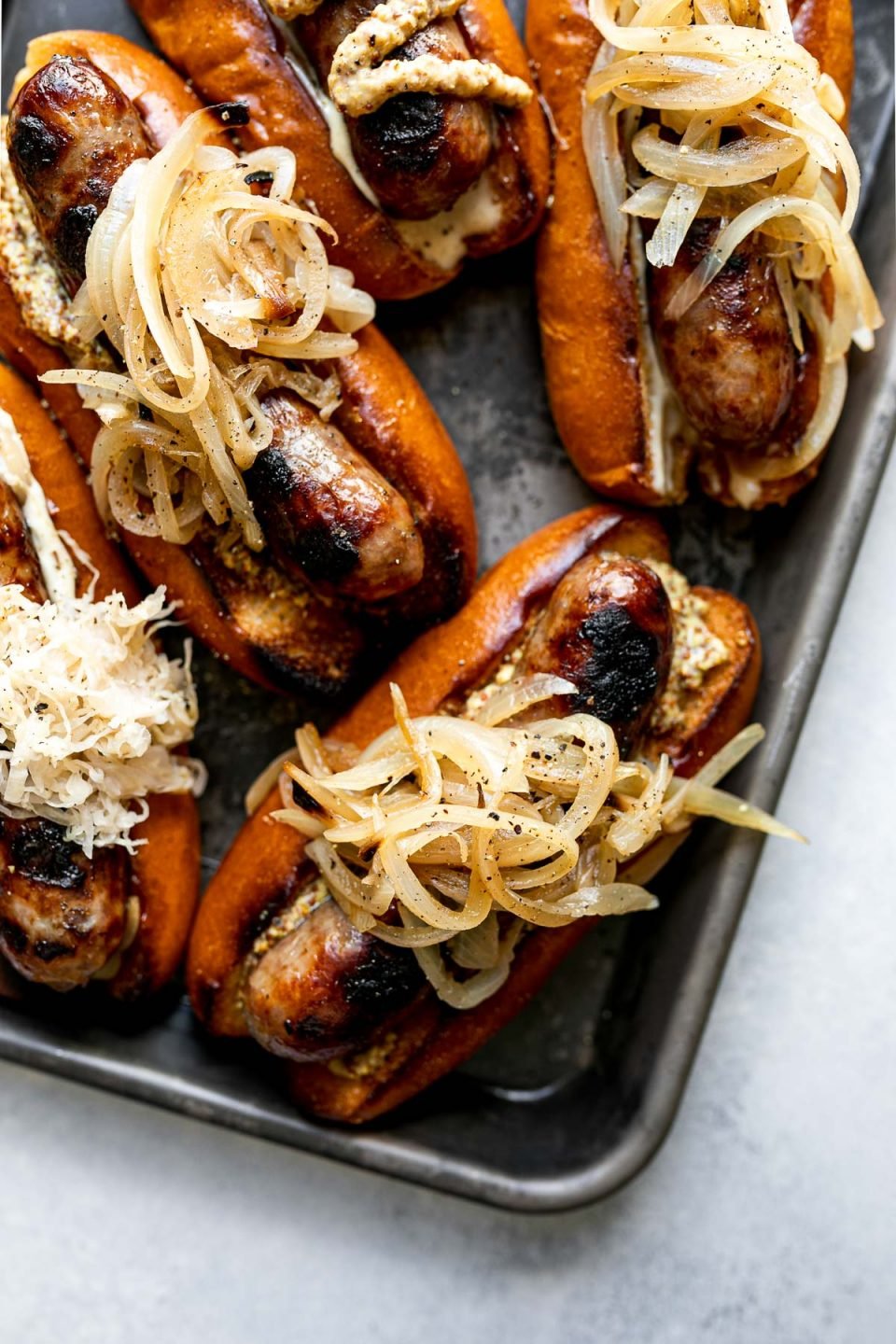 Grilled Wisconsin Beer Brats arranged on a small Nordicware baking sheet. The brats sit on toasted brioche buns, topped with mayonnaise, mustard, braised onions, sauerkraut, etc. The baking sheet sits atop a light blue surface.