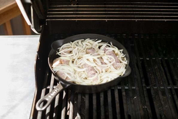 A side angle shot of brats and onions in a black skillet on a grill. 