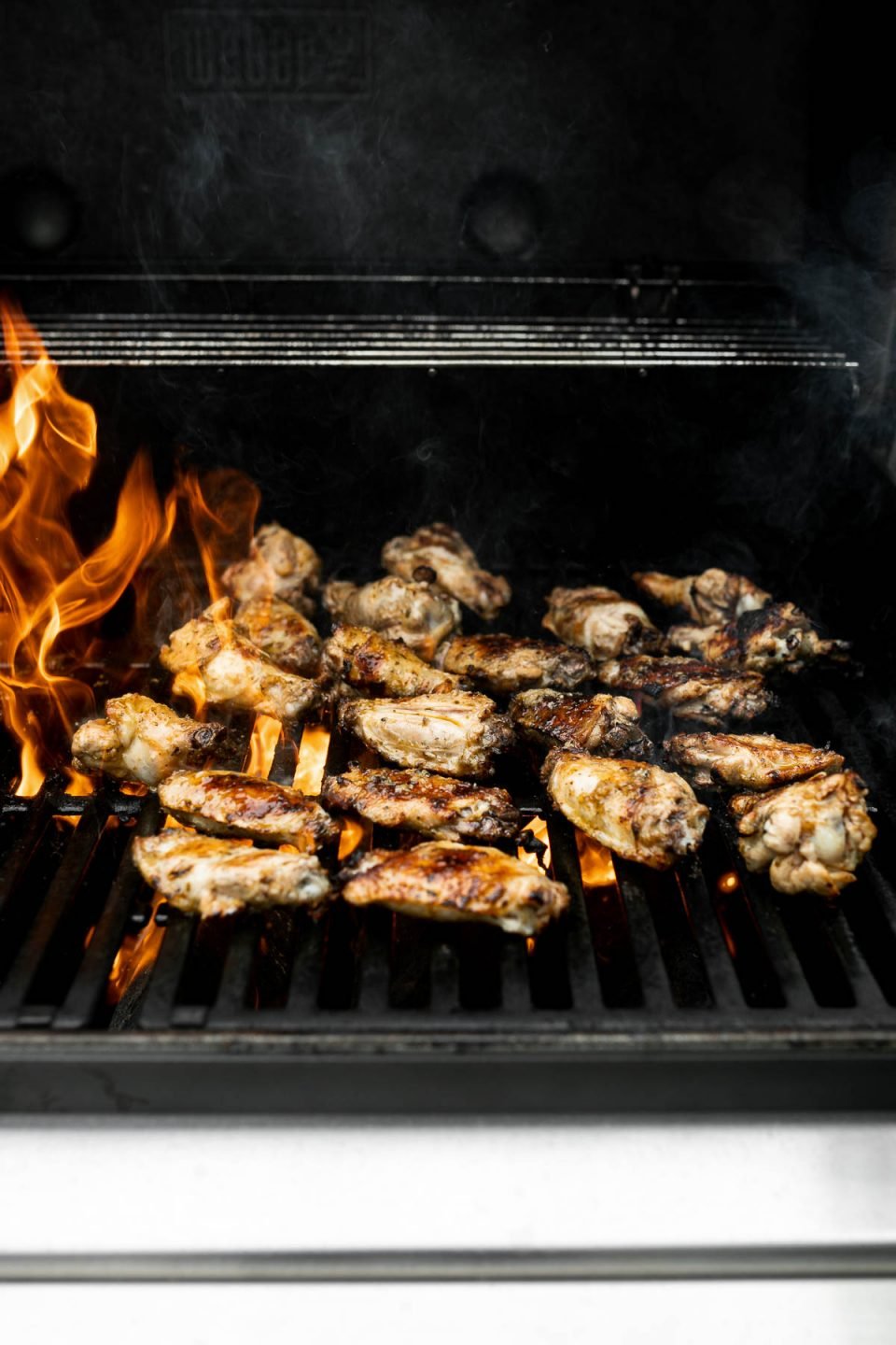 Grilled Greek Chicken Wings being char-grilled on a Weber Grill over direct heat. Flames from the grill burners are present as the chicken wings finish on the grill.