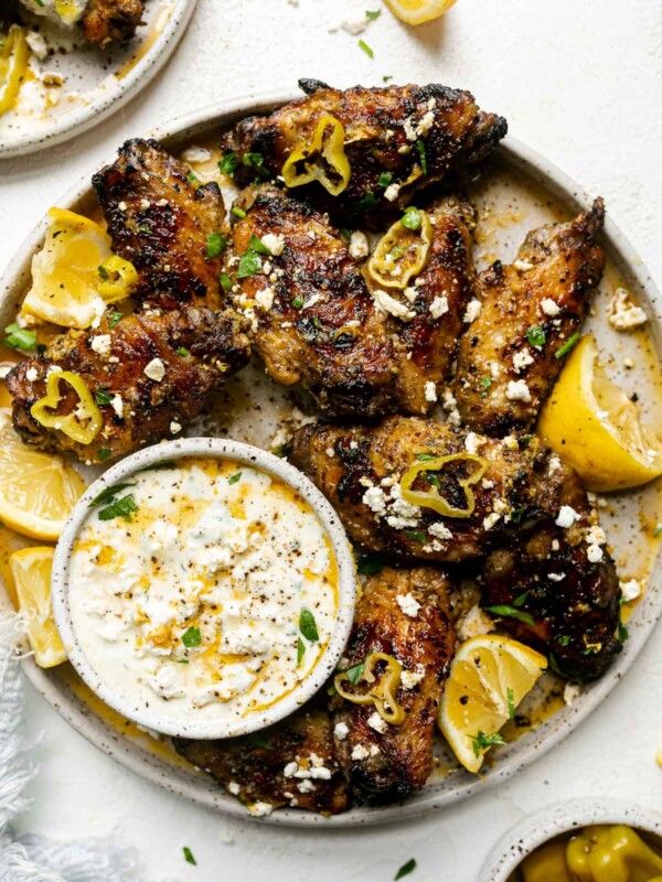 Grilled Greek Chicken Wings arranged on a gray speckled ceramic platter are garnished with herbs, sliced pepperocini, lemon wedges, & feta cheese. A small ceramic bowl is filled with whipped feta dip & also sits on the platter with wings surrounding it. Surrounding the plate is a small plate of wings, a bowl of whole pepperocini peppers, lemon wedges, feta crumbles, loose chopped herbs, & a blue & white striped linen napkin tucked partially under the platter. The platter & surrounding items sit on top of a white textured surface.