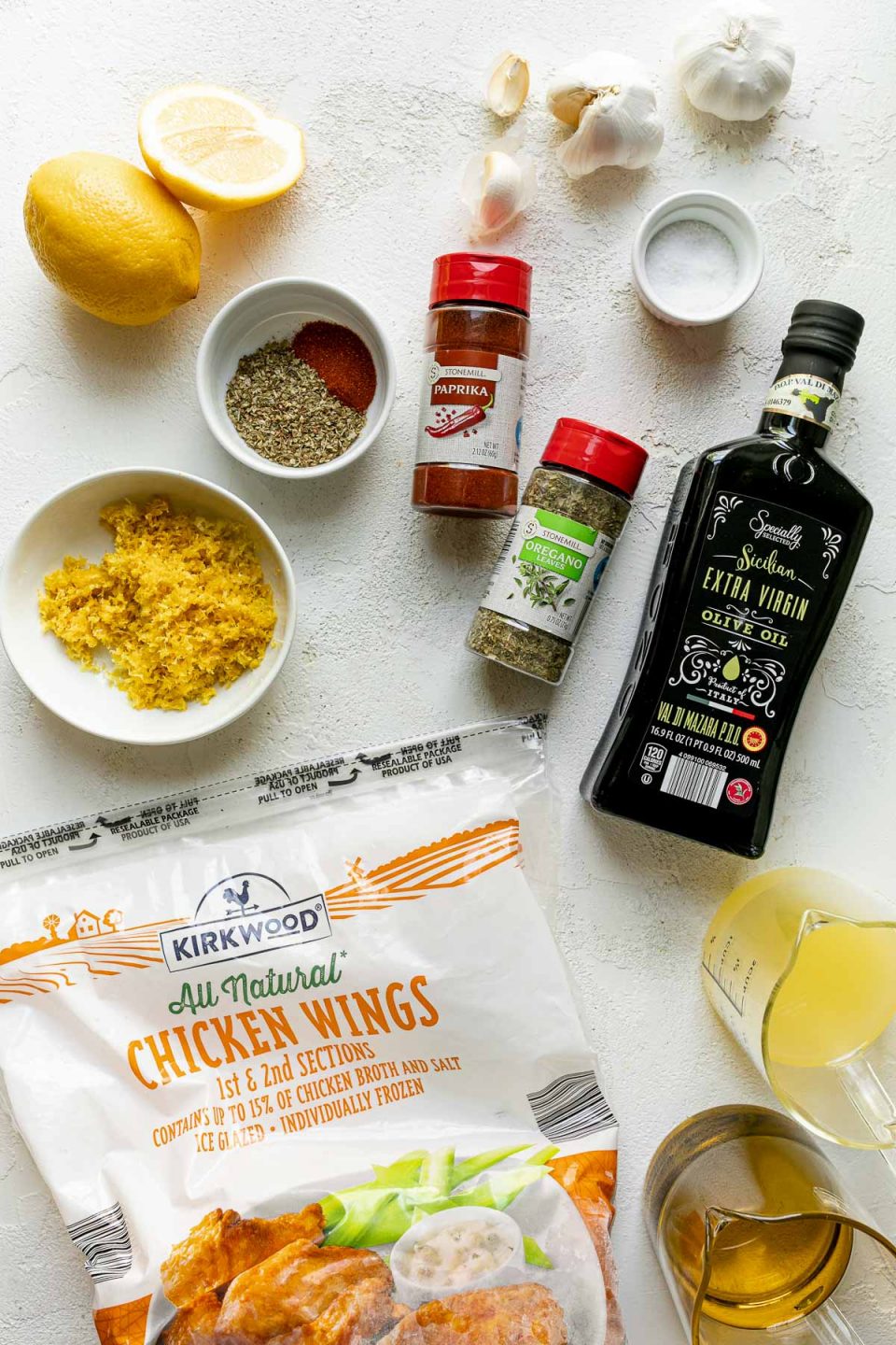 Lemony Greek Grilled Chicken Wings ingredients arranged on a white textured surface – Kirkwood All Natural Chicken Wings from ALDI, kosher salt, Specially Selected Sicilian Extra Virgin Olive Oil from ALDI, lemon zest, lemon juice, garlic, Stonemill Dried Oregano & Paprika from ALDI.
