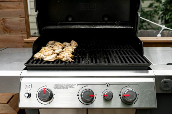 Grilled Lemony Greek Chicken Wings sit on top of Weber Grill grates over indirect heat. The far left grill burner, that the chicken wings sit over is off, while the grill burners to the right are partially turned on to achieve optimum temperature for indirect grilling of the chicken wings.