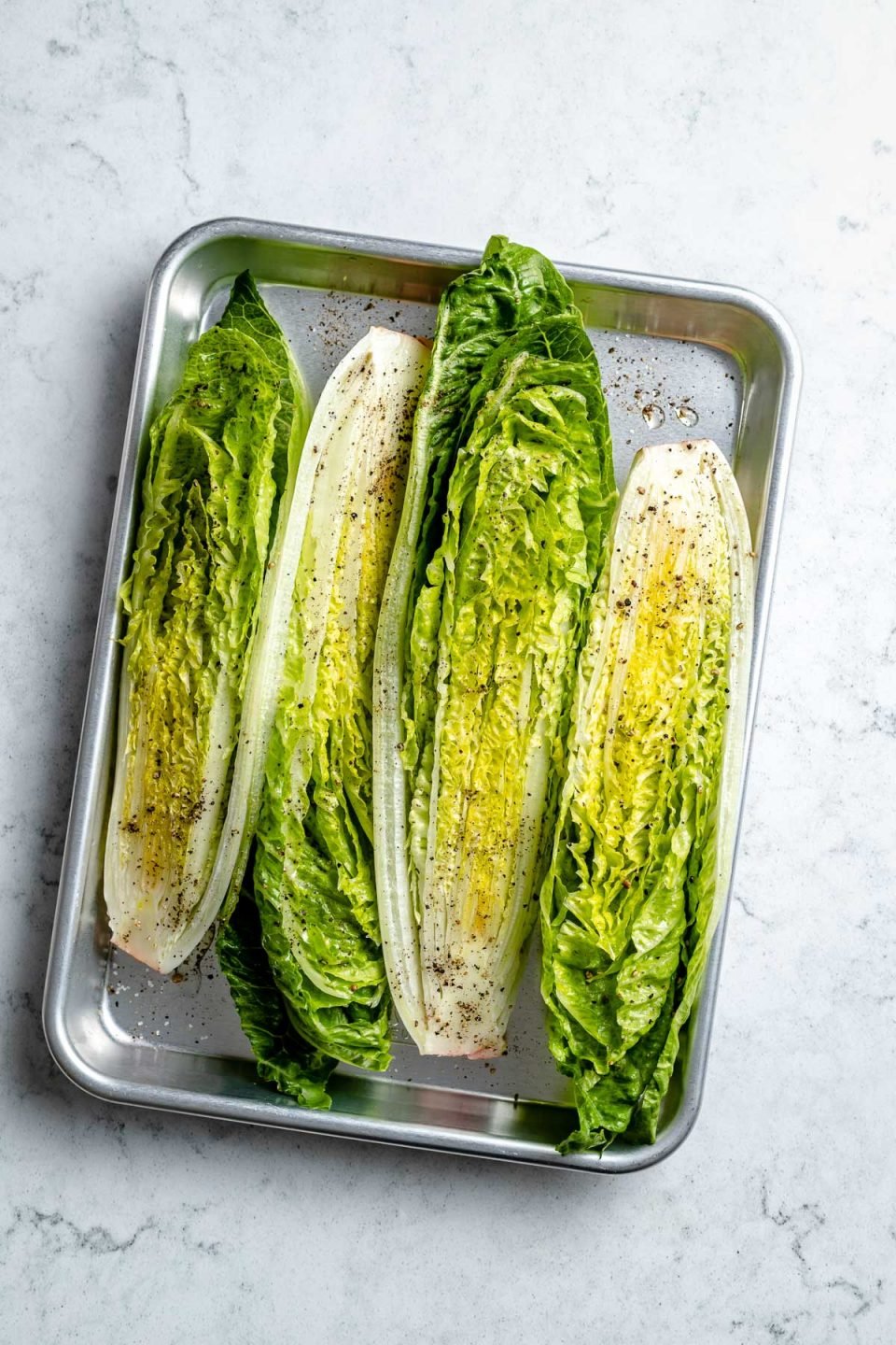 Fresh & uncooked romaine lettuce sliced in half lengthwise & seasoned with oil and ground black pepper lie cut-side up on an aluminum baking sheet. The baking sheet sits on top of a white & gray marble surface.
