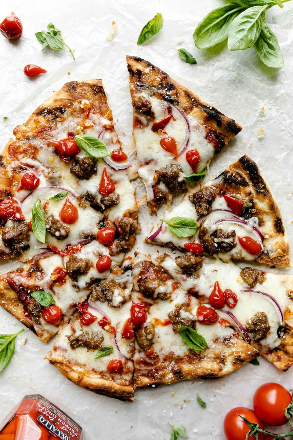 Grilled pizza topped with cheese, Italian sausage, sweetie drop peppers, & fresh basil leaves. The pizza sits atop a white surface, surrounded by fresh basil leaves, tomatoes, & a jar of DeLallo Sweet & Tangy Drop Peppers.