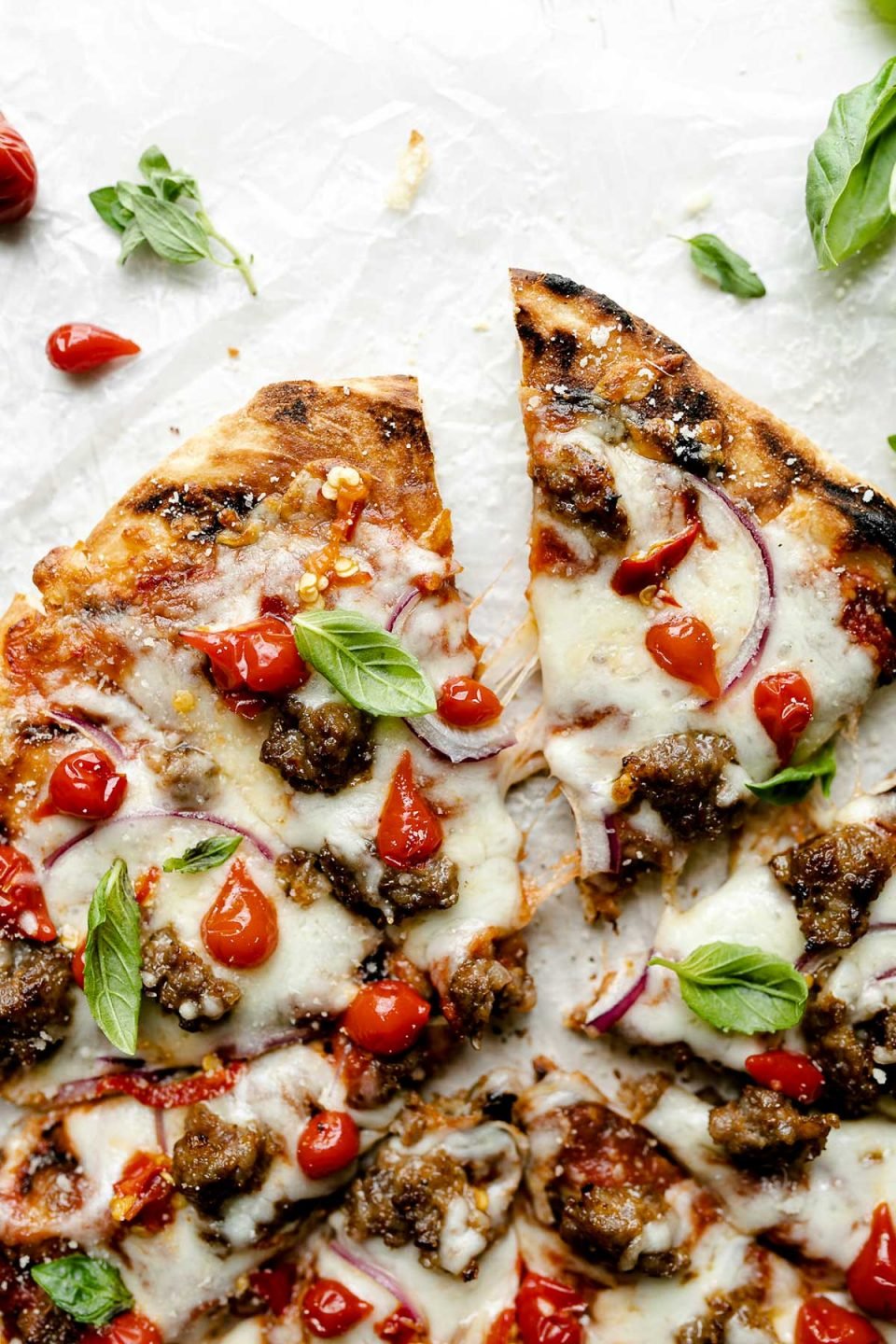 Grilled pizza topped with cheese, Italian sausage, sweetie drop peppers, & fresh basil leaves. The pizza sits atop a white surface, surrounded by fresh basil leaves.