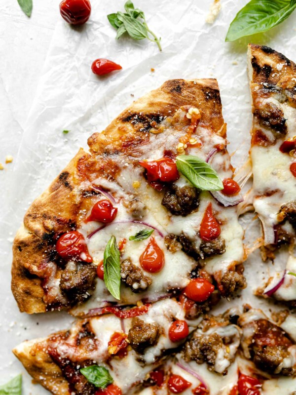 Grilled pizza topped with cheese, Italian sausage, sweetie drop peppers, & fresh basil leaves. The pizza sits atop a white surface, surrounded by fresh basil leaves.