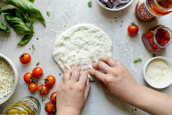 How to grill pizza, Step 3: A woman's hands shown pressing out a fresh ball of pizza dough atop a creamy cement surface, surrounded by fresh basil, cheese, jarred olives, tomatoes, jarred pizza sauce, etc.