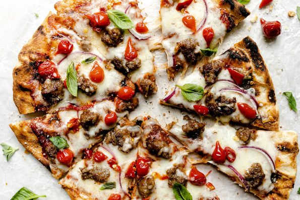 Grilled pizza topped with cheese, Italian sausage, sweetie drop peppers, & fresh basil leaves. The pizza sits atop a white surface, surrounded by fresh basil leaves.