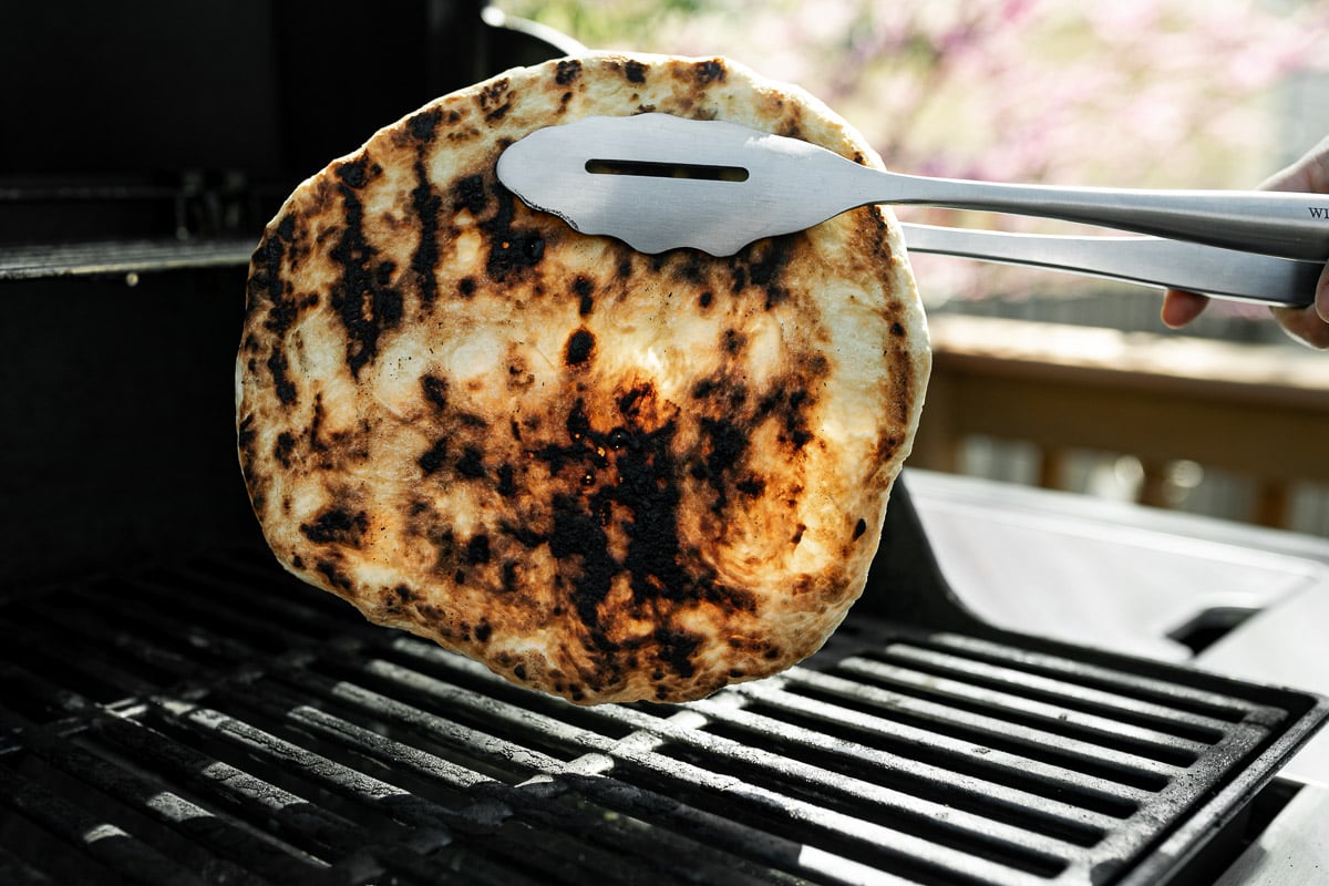 How to grill pizza, Step 4: Grilling tongs holding up par grilled pizza crust over grill grates, revealing perfectly grilled bottom.