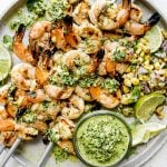 Four zesty grilled shrimp skewers with jalapeño pesto & grilled corn salsa stacked on top of a gray speckled ceramic plate & garnished with lime wedges. A small jar of jalapeño pesto also sits on the plate ready for dipping or drizzling. The plate sits atop a light gray & white surface.
