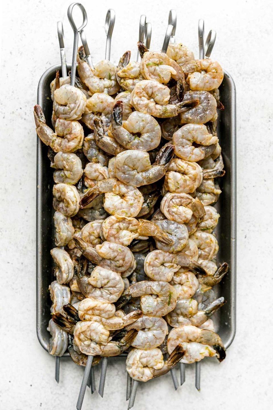 Multiple skewers of zesty marinated jumbo shrimp are stacked on top of an aluminum baking sheet. The baking sheet sits on top of a white & light gray textured surface.