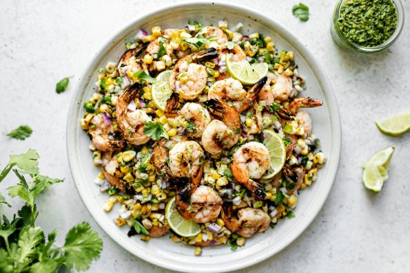 Zesty Grilled Shrimp with Jalapeño pesto & grilled corn salsa on top of a white ceramic plate & garnished with lime wedges. The plate sits atop a light gray & white surface, next to a bunch of cilantro, a few loose cilantro leaves, a small jar of jalapeño pesto, & a couple of squeezed lime wedges.