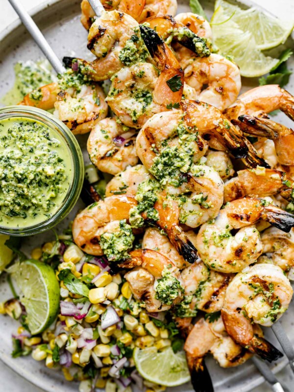 A close up of four zesty grilled shrimp skewers with jalapeño pesto & grilled corn salsa stacked on top of a gray speckled ceramic plate & garnished with lime wedges. A small jar of jalapeño pesto also sits on the plate ready for dipping or drizzling. The plate sits atop a light gray & white surface.