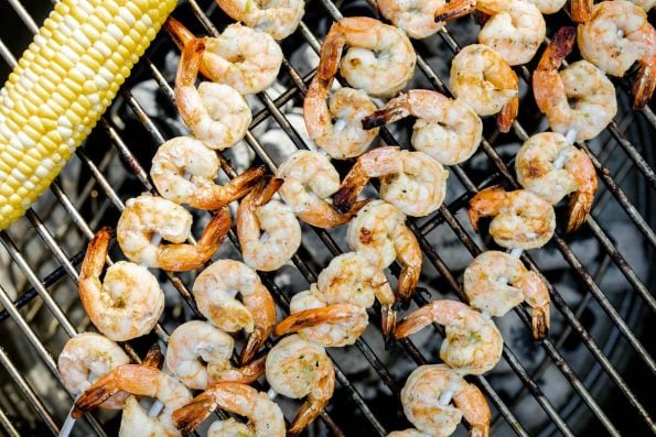 A close up of four grilled shrimp skewers and a cob of sweet corn grilling on top of a Solo Stove Grill. The grilled shrimp are becoming opaque & developing a golden brown char from the grill.